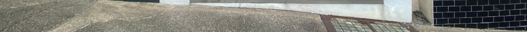 A low-resolution, ground-level photograph showing a paved sidewalk, a metal drain grate, and the base of a black tiled wall.
