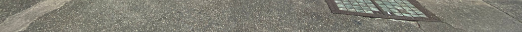 A close-up photograph showing a section of asphalt pavement with a metal drainage grate visible in the upper right corner.
