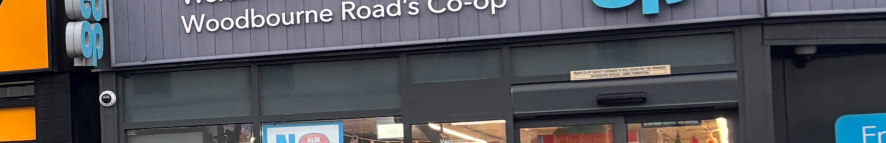 A close-up photograph of the exterior shopfront of a Co-op store on Woodbourne Road, showing the dark fascia and large glass windows.