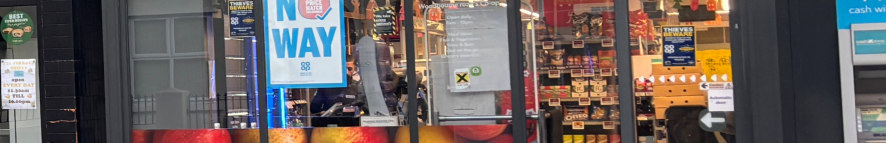 A street-level photograph showing the exterior of a commercial shop front with large glass windows and doors. The image displays the existing facade including signage and interior shelving visible through the glass.