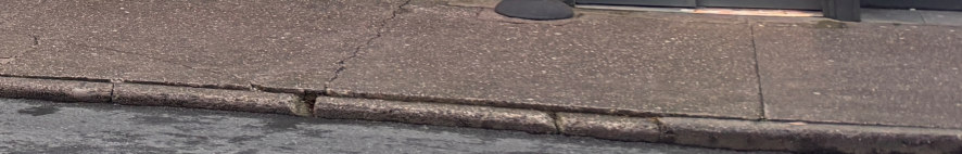 A close-up photograph showing a cracked concrete pavement and curb with a manhole cover visible at the top.