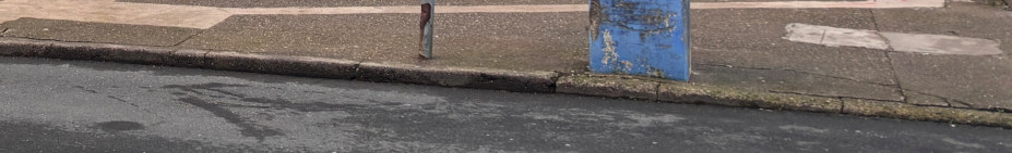 A low-angle photograph showing a paved sidewalk, a concrete curb, and an asphalt road surface with a blue utility box or bollard base.