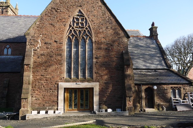 A photograph showing the exterior brick facade of a historic building, likely a church, featuring a large Gothic window above a wooden door with white blocks on the ground.