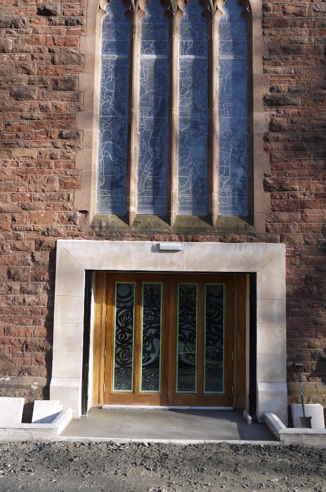 A photograph of a brick building exterior featuring a large stained glass window above a wooden double door with decorative glass panels.