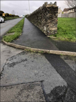 A photograph showing a paved path or driveway running alongside a road, bordered by a tall stone wall on the right.