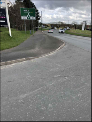 A photograph showing a paved road curving towards a roundabout with a green directional sign on the left and vehicles in the distance.