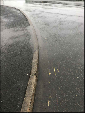 A close-up photograph showing a wet asphalt road surface, a concrete curb, and faint yellow painted markings on the ground.