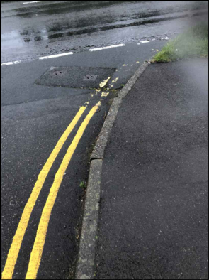 A close-up photograph of a wet asphalt road surface featuring double yellow lines along the curb and a utility cover.