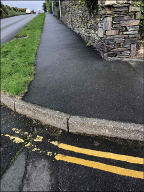 A photograph showing a paved footpath running alongside a road, bordered by a stone wall on the right and a grass verge on the left.