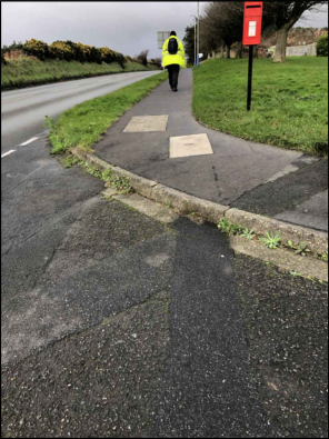 A photograph showing a paved footpath running alongside a road, with a person in high-visibility clothing walking away and a red post box on the grass verge.