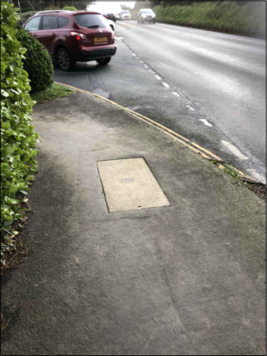 A photograph showing a paved footpath or driveway entrance next to a road, featuring a rectangular concrete slab and a red car in the background.