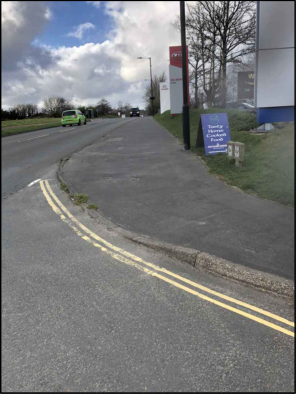 A street-level photograph showing a road with double yellow lines curving past a commercial building and a blue planning sign.