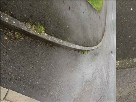 A close-up photograph showing a curved concrete curb and asphalt surface with some moss growth.