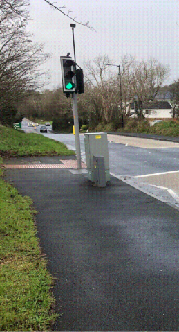 A photograph showing a pedestrian crossing signal with a green light and a control box on a paved path beside a road.