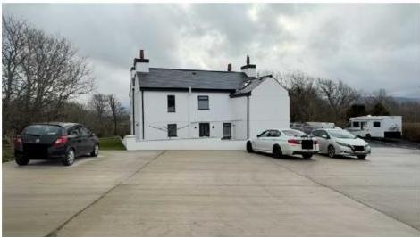 A photograph showing a white, two-story building with a slate roof and a large concrete driveway in a rural setting.