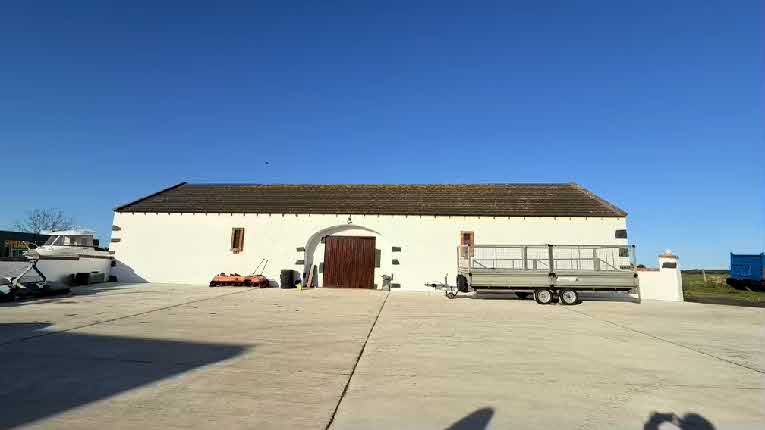 A photograph showing a large white agricultural building with a tiled roof and a central arched wooden door. A concrete yard is in the foreground with a boat trailer on the left and a livestock trailer on the right.
