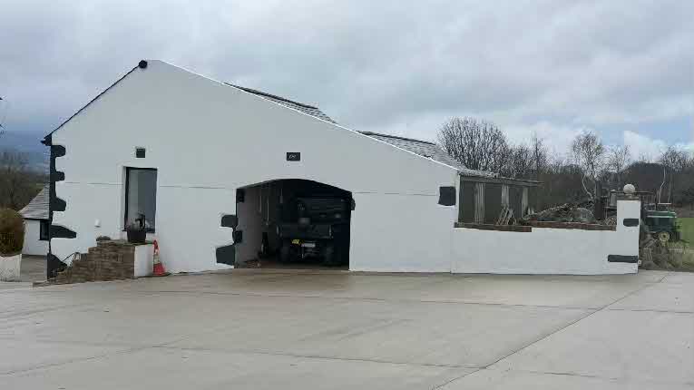 A photograph showing a modern white agricultural building with a large arched opening housing a vehicle, set against a rural backdrop.