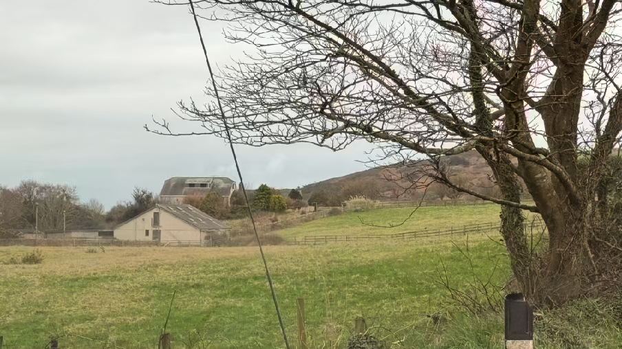 A rural landscape photograph showing a grassy field with existing agricultural buildings and a stone structure in the background.