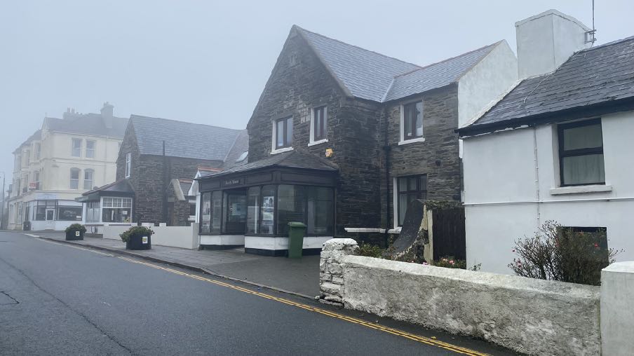 A street-level photograph taken on a foggy day showing a row of buildings, including a dark stone building with a shop front and a white residential property.