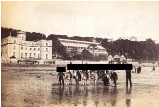 A vintage sepia-toned photograph depicting a coastal scene with large buildings, likely a hotel, and people walking on the beach.