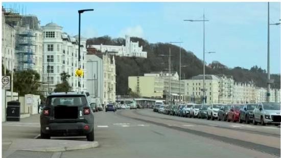 A street scene in Douglas showing a black car in the foreground, a row of parked cars, and large white hotel buildings along the coast with a castle on a hill in the background.