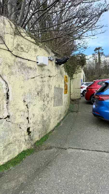 A photograph showing a weathered stone wall with a mounted projector unit and electrical box, adjacent to a car park with parked cars.
