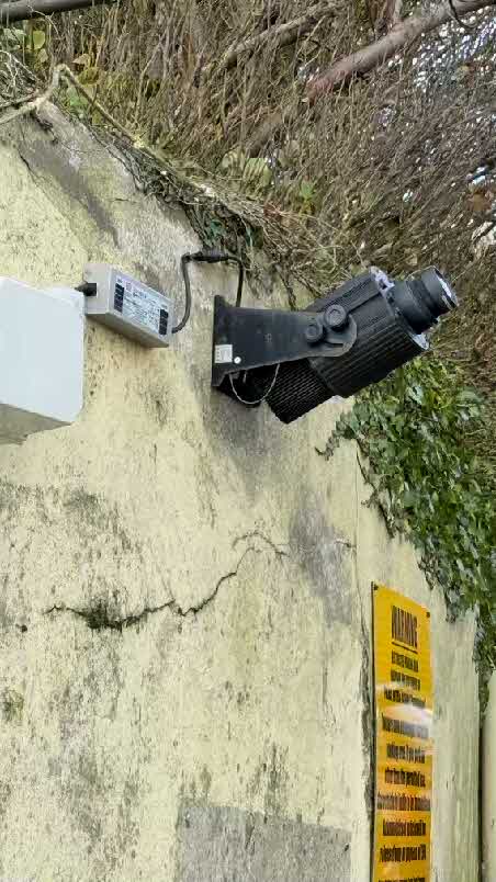 A close-up photograph showing a large black projector and power unit mounted on a textured exterior wall with vegetation growing above.