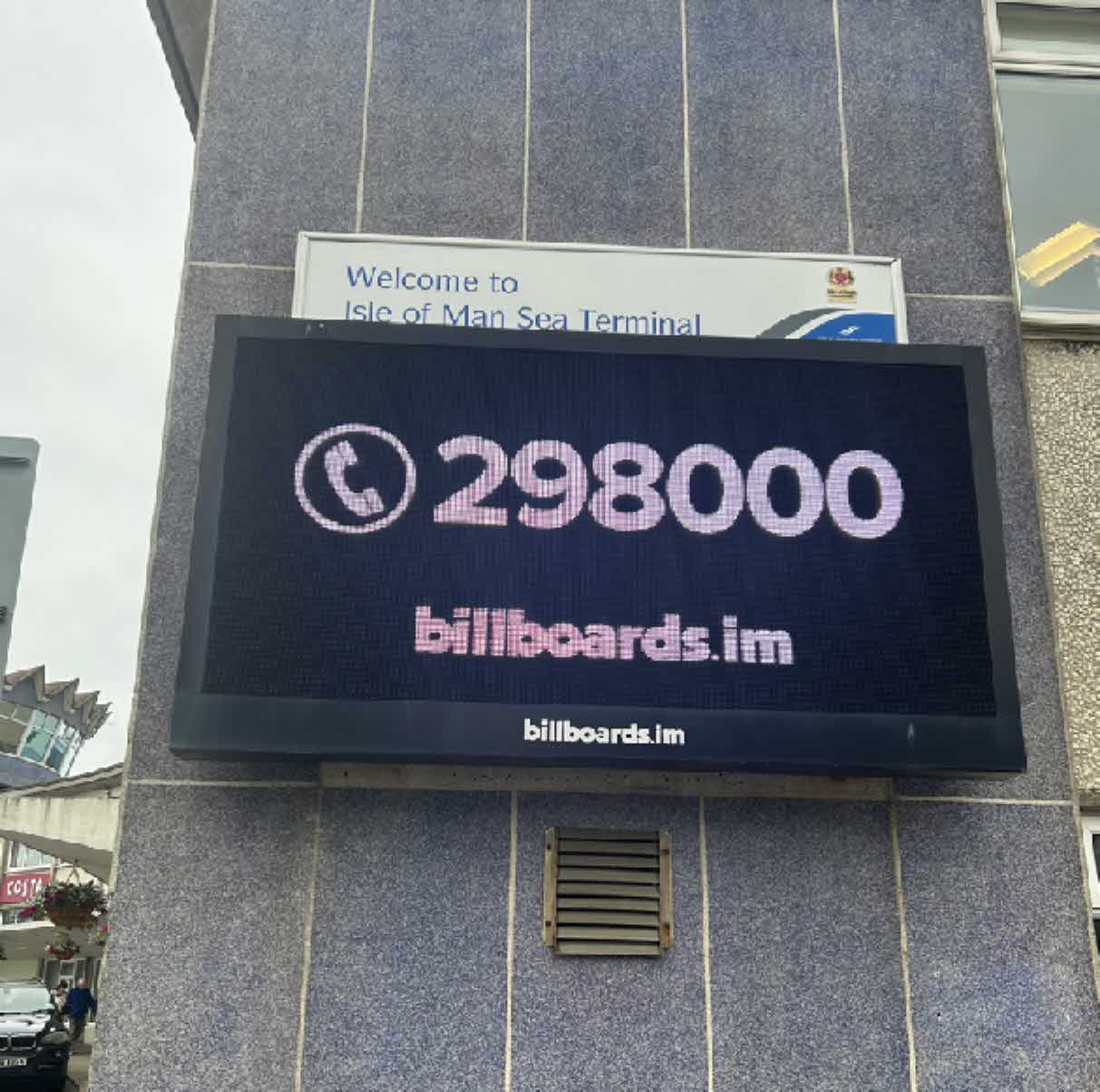 A photograph showing a large digital LED sign mounted on a grey building facade, displaying a phone number and website, situated below a 'Welcome to Isle of Man Sea Terminal' sign.