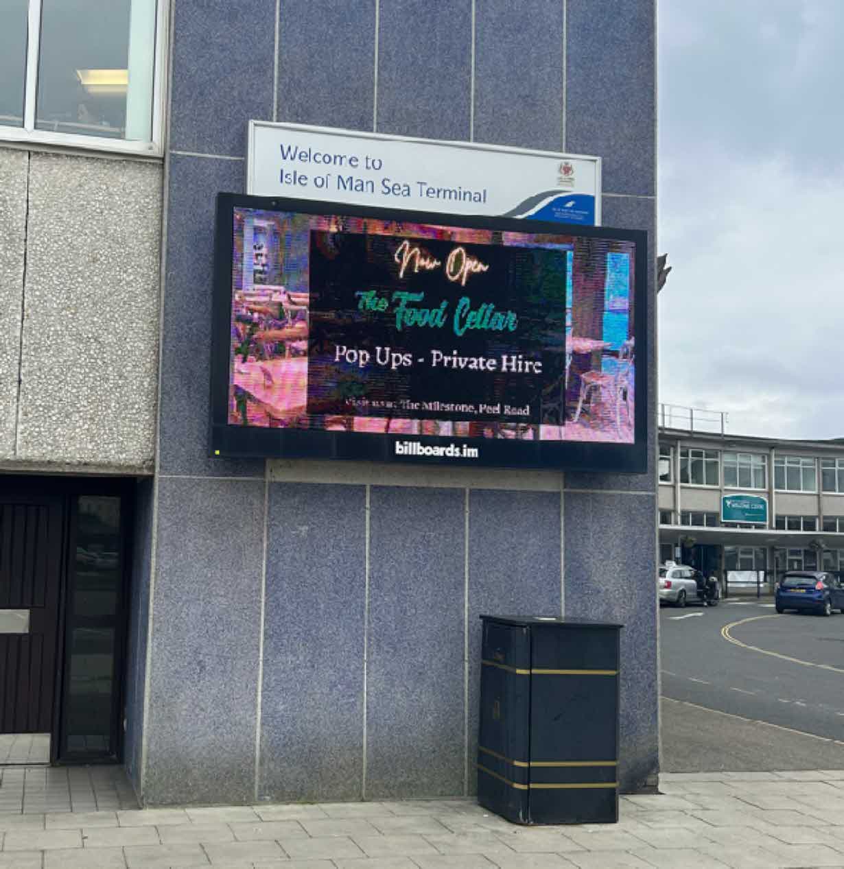 A photograph showing a large digital advertising screen mounted on the grey exterior wall of the Isle of Man Sea Terminal building.
