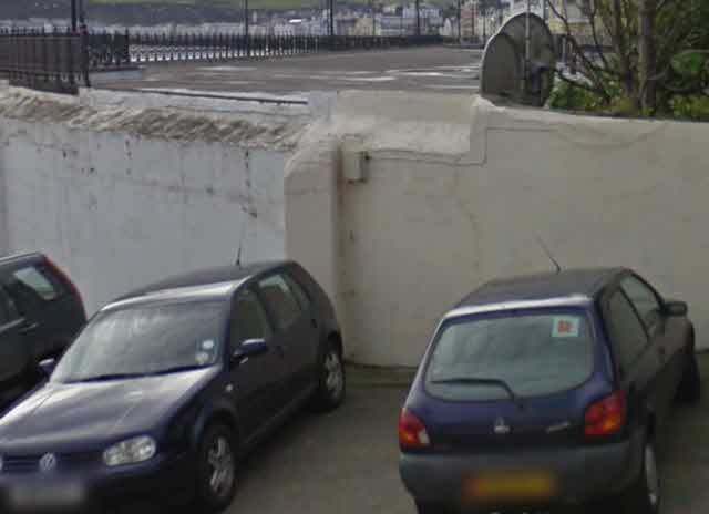 A photograph showing a white boundary wall in a car park with parked cars in the foreground and a coastal promenade in the background.