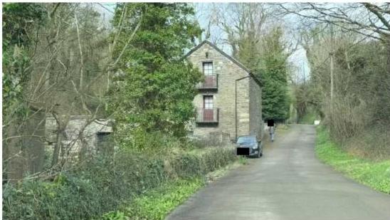 A photograph showing a two-story stone building situated beside a narrow rural road, with a car parked in front and surrounding vegetation.