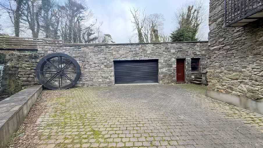 A photograph showing the exterior of a stone building featuring a large water wheel, roller shutter door, and paved courtyard.