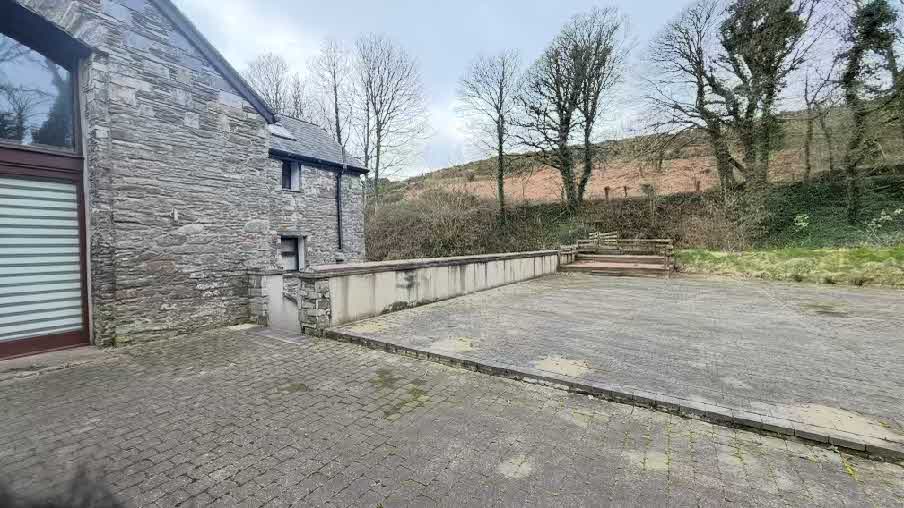 Exterior photograph of a stone building with a large paved driveway and a low stone wall bordering a grassy hillside.