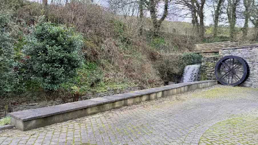 A photograph of a paved outdoor area featuring a low stone wall, a small waterfall, and a large wooden water wheel. The background shows a steep, wooded bank.
