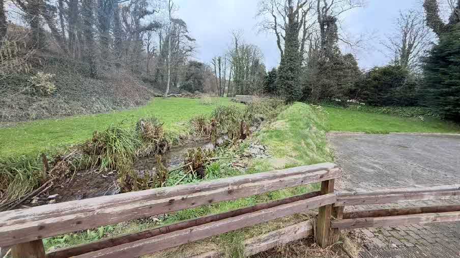 A photograph showing a rural site with a wooden fence in the foreground, a small stream or ditch, and a paved driveway area surrounded by trees and grassy banks.