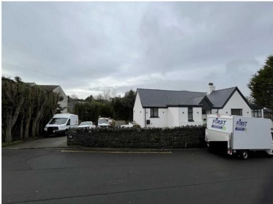 A street-level photograph showing a white detached house with a grey roof, fronted by a stone wall and a driveway with parked white vans.