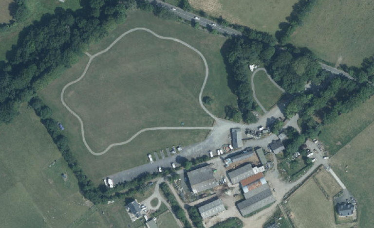 An aerial view of a rural site showing a large grassy field with a proposed light-colored track layout looped around the perimeter, adjacent to existing buildings.