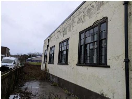 A photograph showing the weathered side elevation of a white rendered building with black sash windows and a downpipe, taken from a wet alleyway.