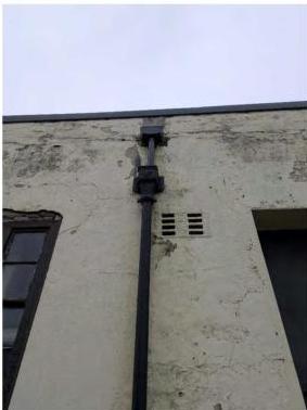 A low-angle photograph showing the weathered exterior wall of a building, featuring a black drainpipe and ventilation grilles.