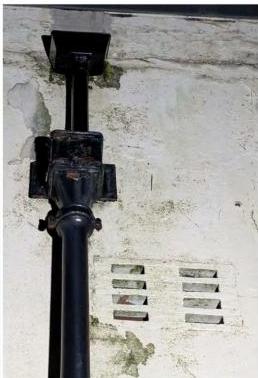 A close-up photograph showing a black vertical drainpipe attached to a weathered white wall with a ventilation grille nearby.
