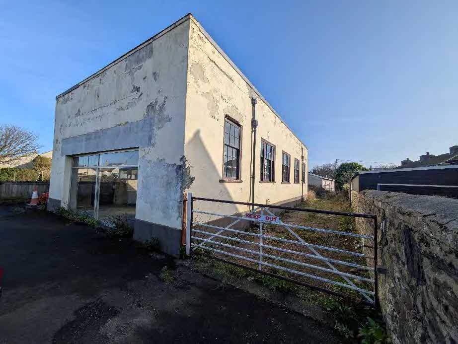 A photograph showing a dilapidated, single-story white building with peeling paint and large windows, viewed from the side with a metal gate in the foreground.
