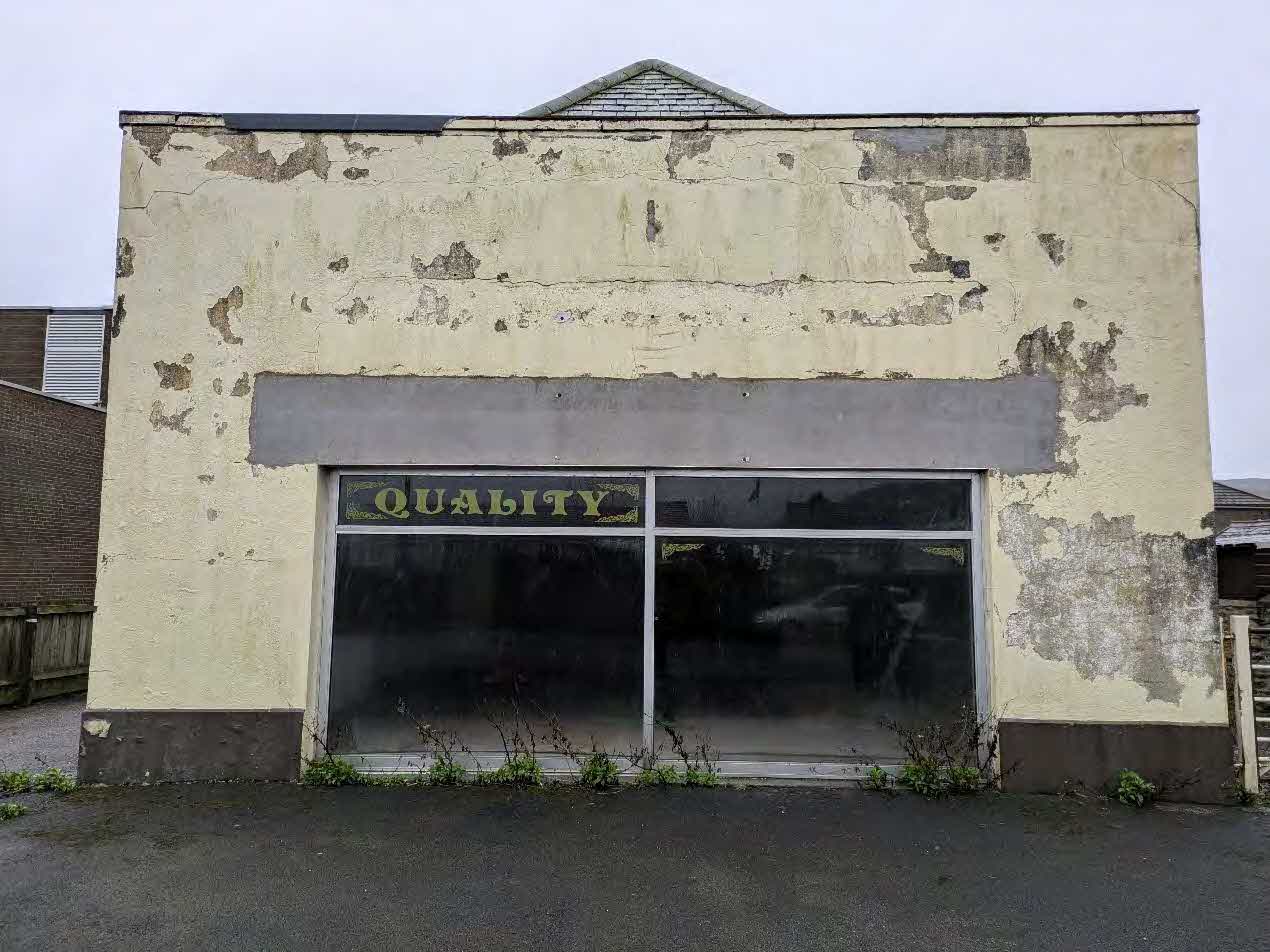 A photograph showing the weathered exterior facade of a single-story commercial building with peeling render and a large storefront window.