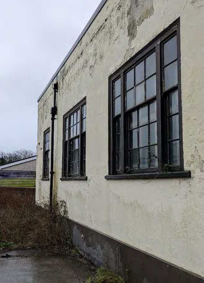 A photograph showing the weathered cream exterior wall of a building featuring dark-framed multi-paned windows and a vertical drainpipe.