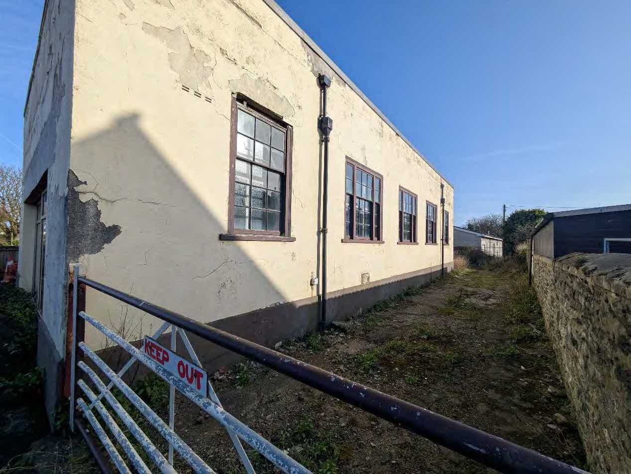 A photograph showing the side elevation of a weathered, single-story building with peeling render and multi-pane windows, likely an outbuilding or former agricultural structure.