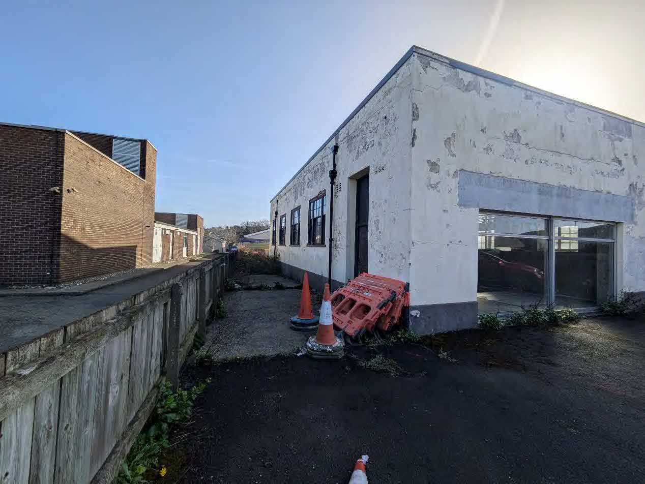 A photograph showing a weathered white single-story building with large glass windows, likely a garage or workshop, situated next to a brick building and wooden fence.