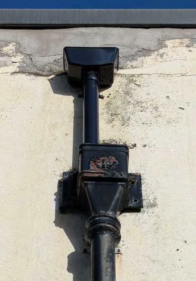 A close-up photograph showing a black metal rainwater downpipe and drainage fitting attached to a weathered, light-colored exterior wall.