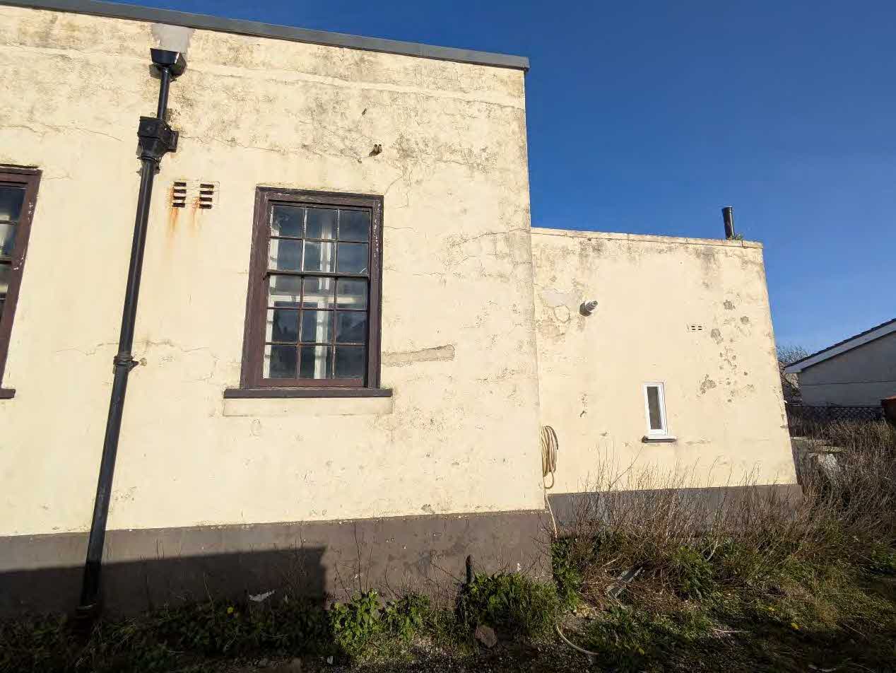 Photograph showing the exterior wall of a cream-rendered building with a multi-paned window and a black drainpipe. The ground in the foreground is overgrown with vegetation.