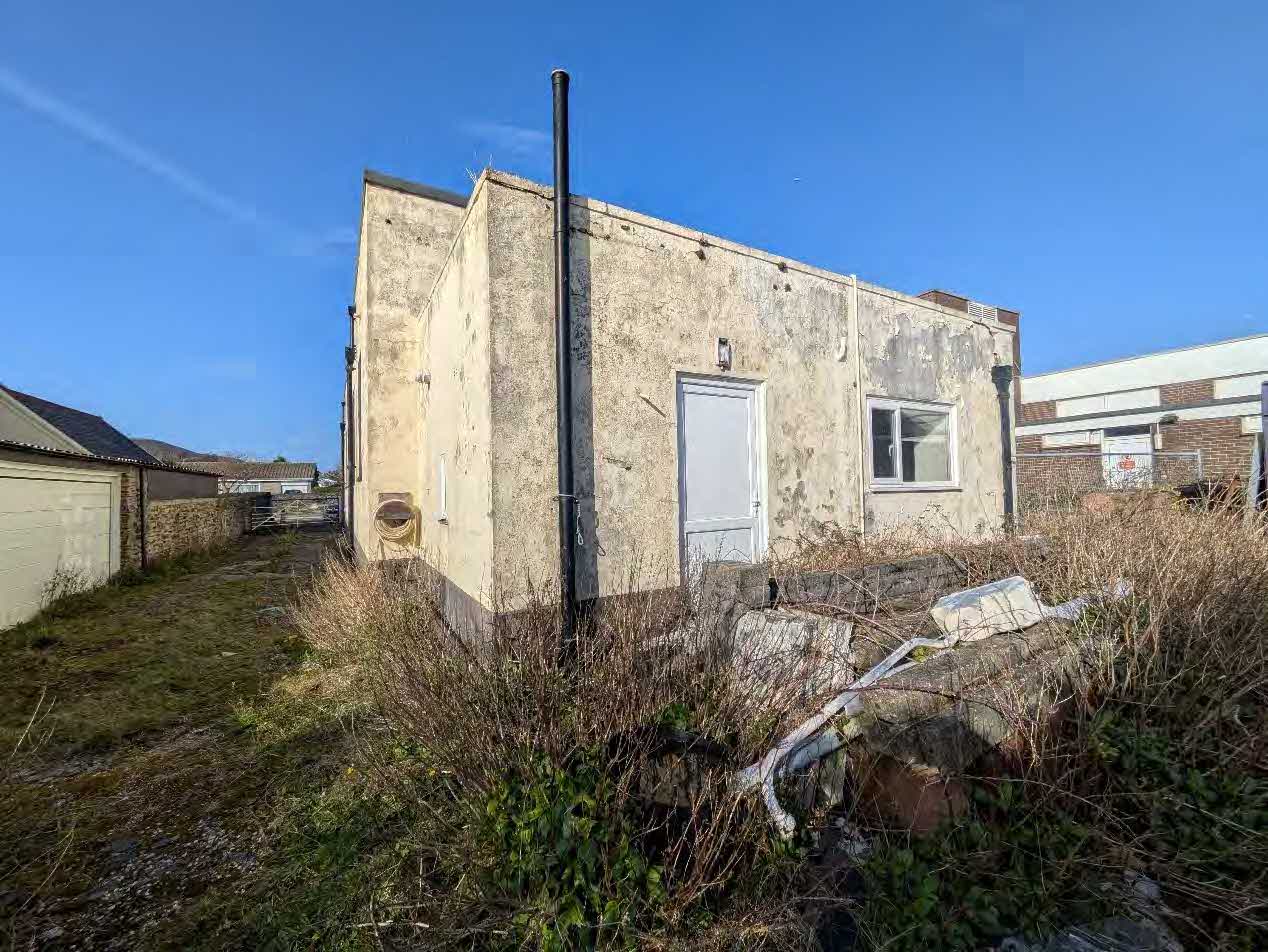 A photograph showing a weathered, single-story building with peeling render and an overgrown front garden area.