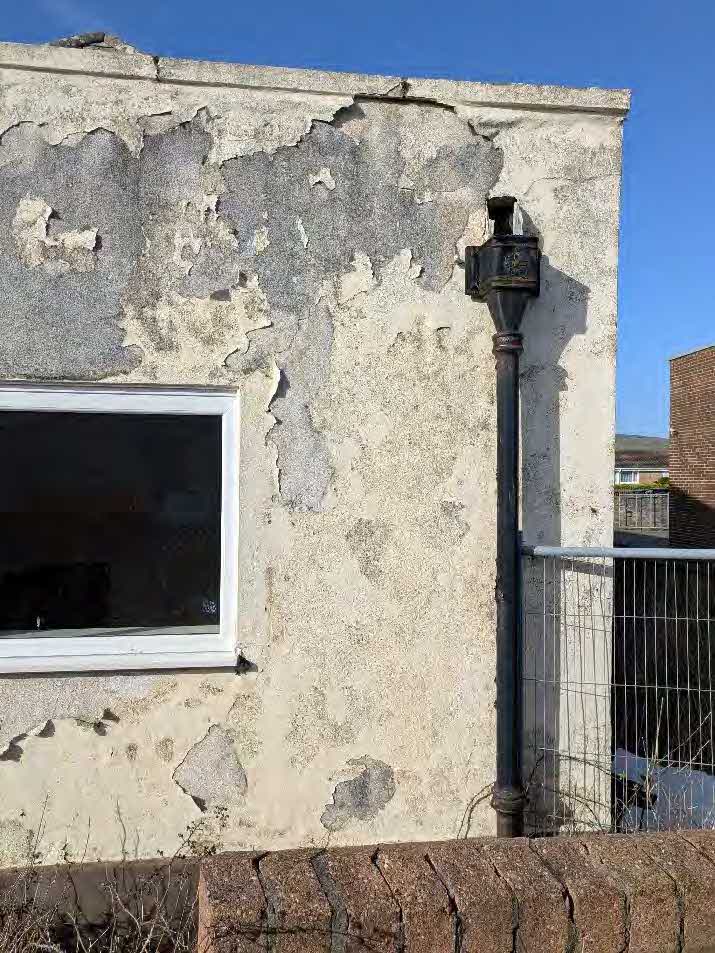 A close-up photograph of a weathered building exterior showing significant peeling render, a white window frame, and a black drainpipe.