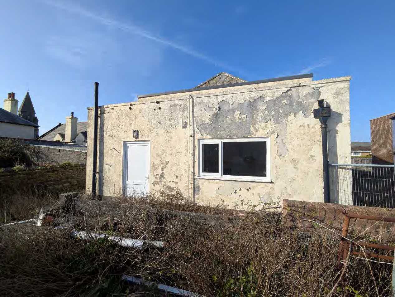 A photograph showing the exterior of a weathered, single-story building with significant peeling render and overgrown vegetation in the foreground.